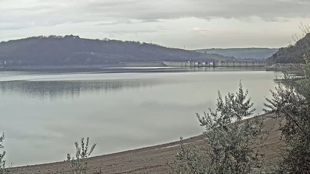 A calm lake with a dam and low hills in the background under an overcast sky.