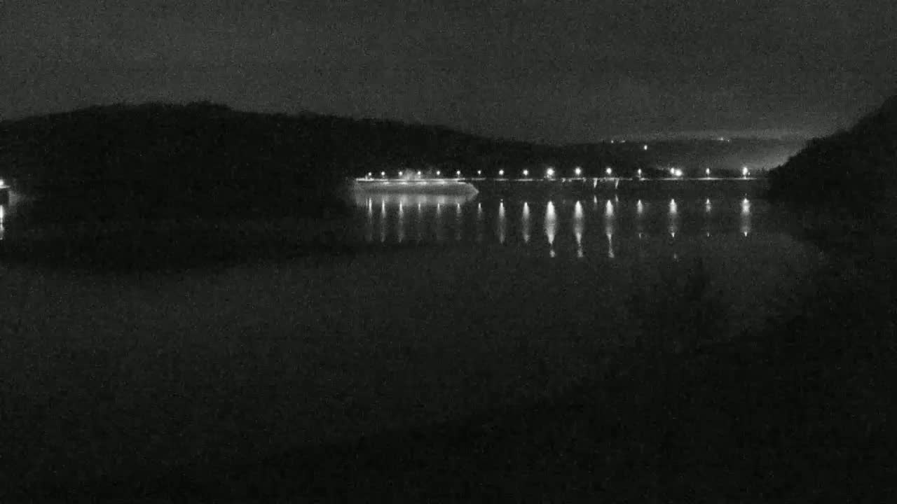 A dark, nighttime scene shows a body of water reflecting the lights of a structure on the far shore, under a clear, moonless sky.