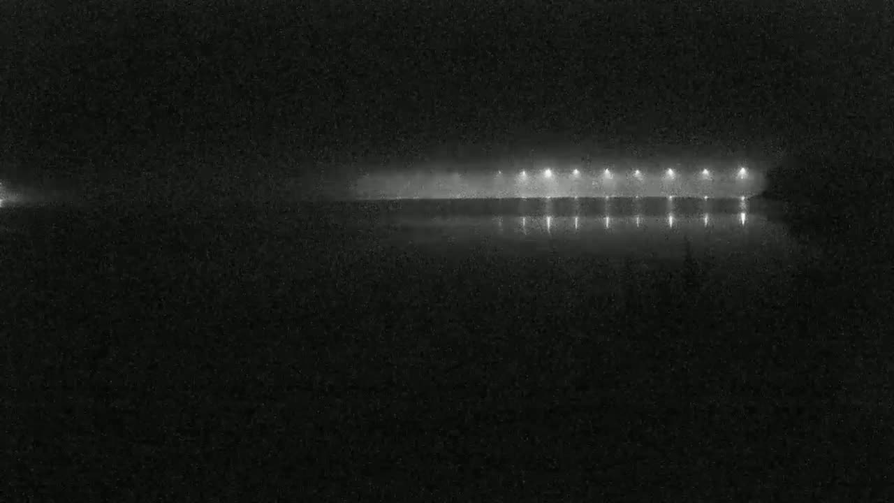 A foggy night scene shows a long line of lights reflected in a dark body of water.
