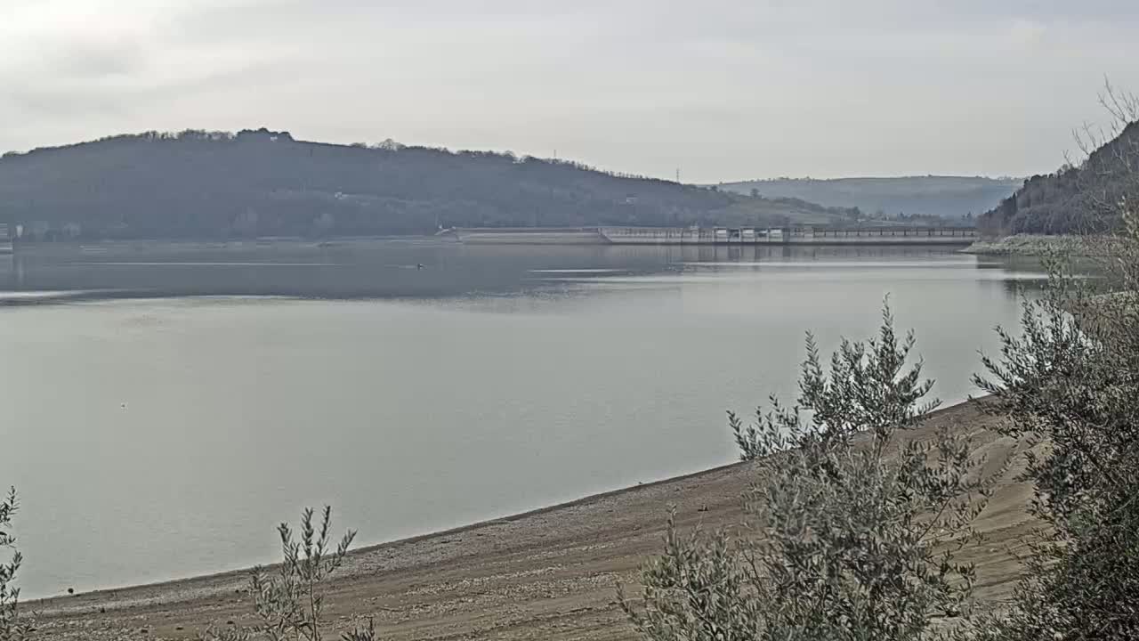 A calm lake with a dam in the background, under an overcast sky, shows a receding shoreline with exposed, dry earth.