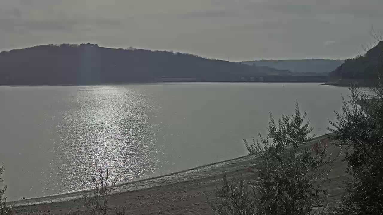 A calm lake reflects sunlight under an overcast sky, with hills visible in the distance and shrubs in the foreground.
