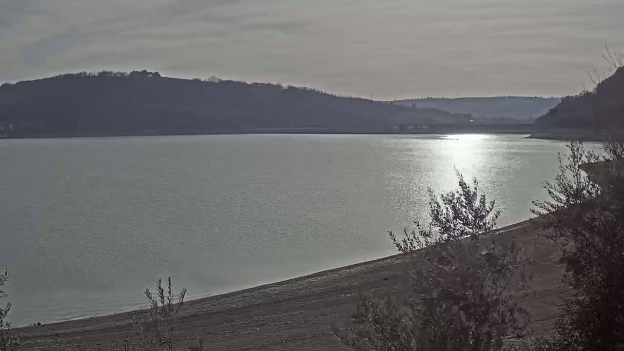 A calm lake reflects sunlight, its shore lined with sparse, low-lying vegetation under a mostly cloudy sky.