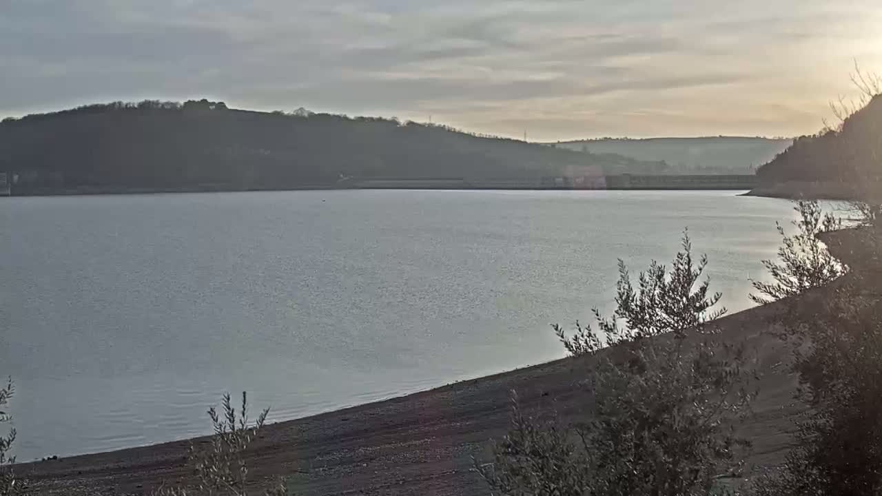 A calm lake stretches out beneath a partly cloudy sky, nestled between gently sloping hills.