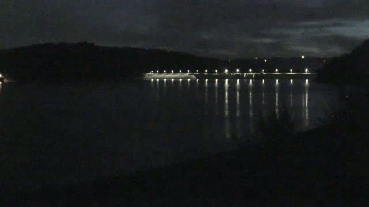 A dark, nighttime scene shows a body of water reflecting the lights of a dam across its surface under a partly cloudy sky.