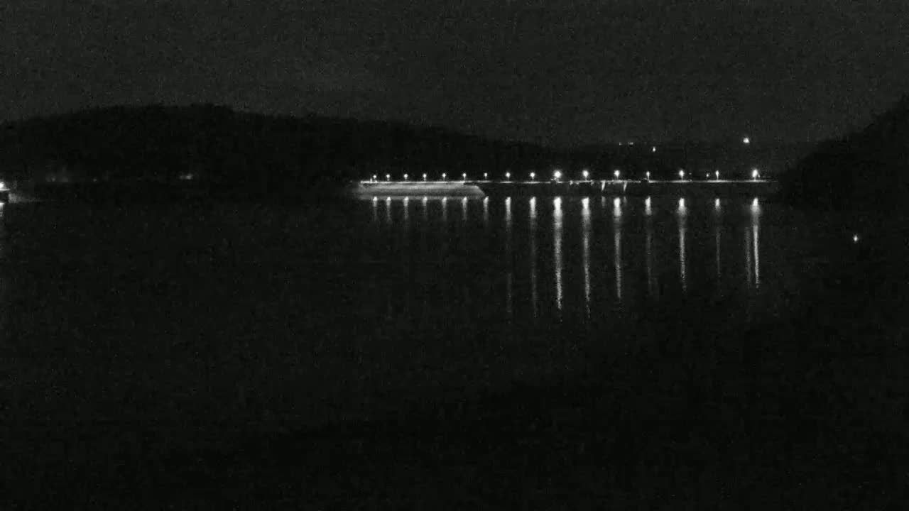 At night, a long, illuminated structure stretches across a dark body of water, reflected lights shimmering on the still surface.