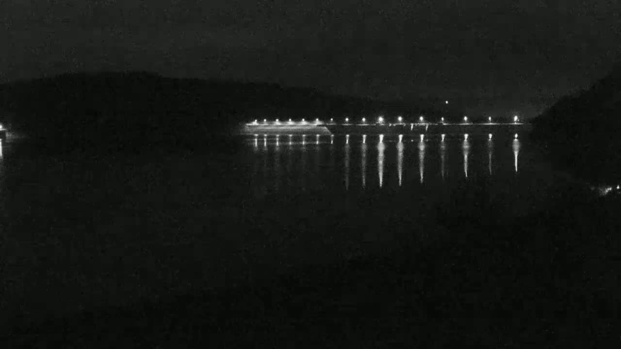 At night, a long, low-lit structure is visible across a dark body of water, its lights reflecting on the still surface.