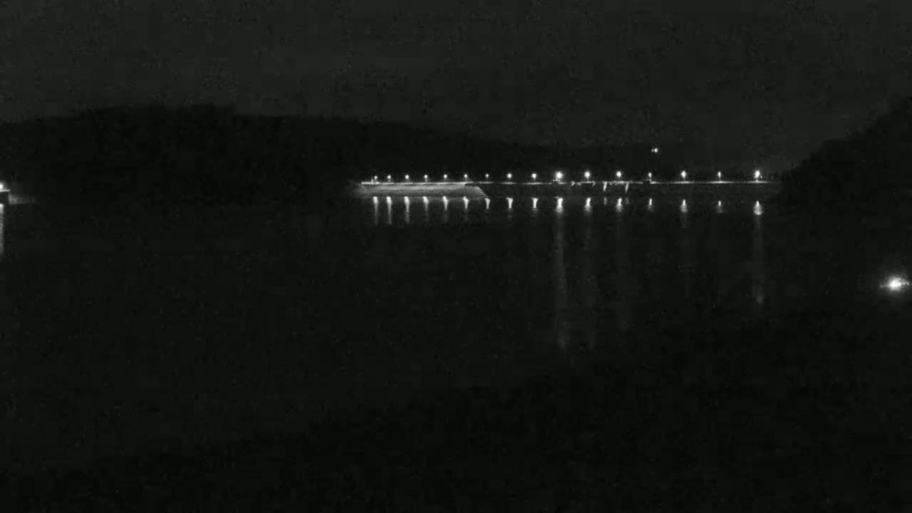 At night, a long, dark structure with lights is visible across a body of dark water.