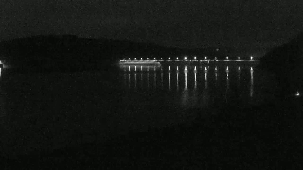 At night, a long, lit structure stretches across a dark body of water, reflecting its lights onto the still surface.
