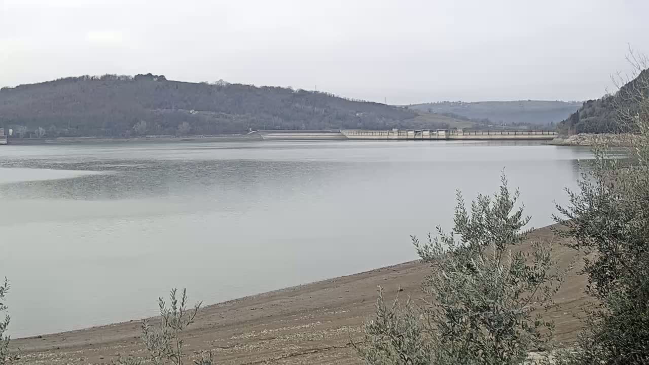 A calm, gray day reveals a reservoir with low water levels, exposing a wide expanse of dry, sandy shoreline, backed by a dam and tree-covered hills.
