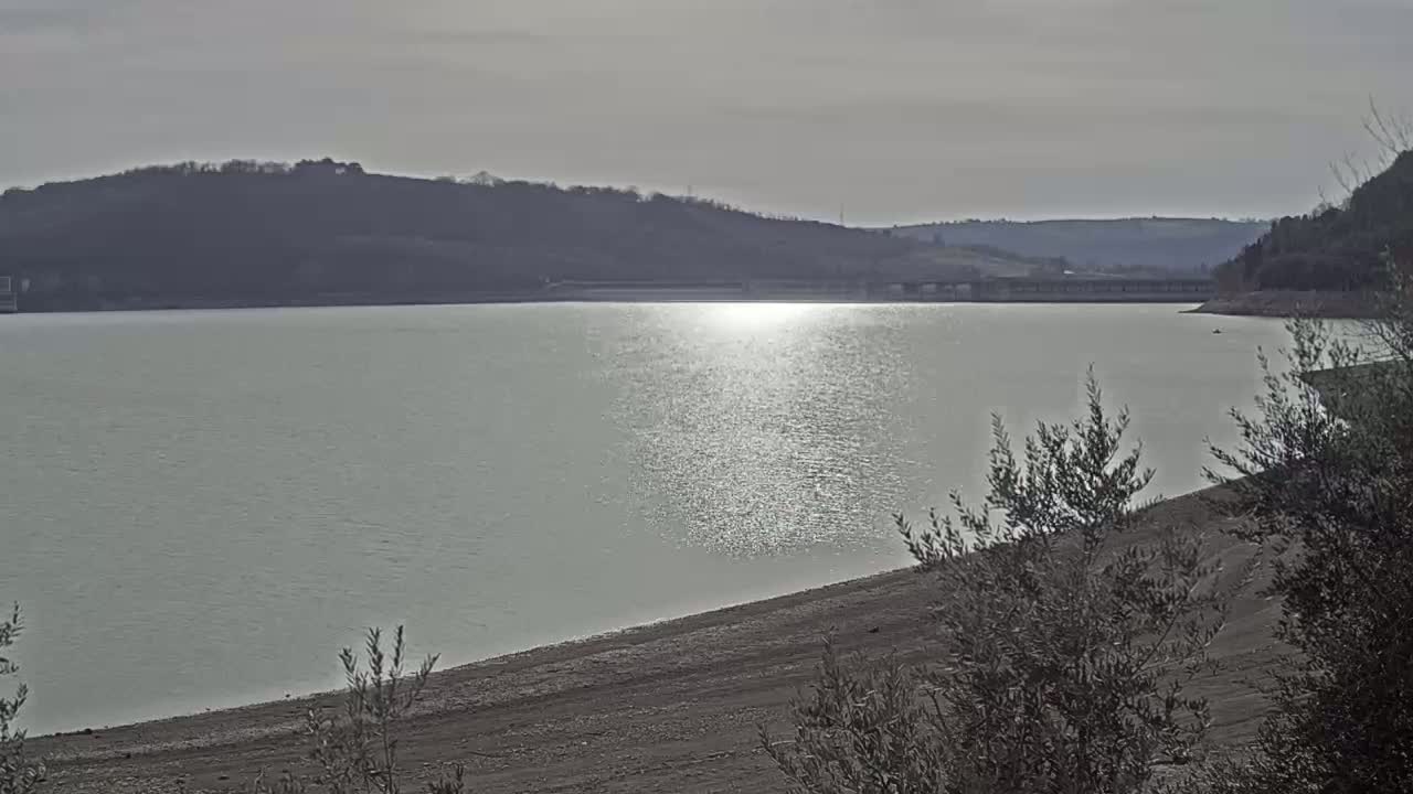 A calm, light-green lake reflects sunlight under a mostly cloudy sky, with hills in the background and sparse, low-lying vegetation in the foreground.