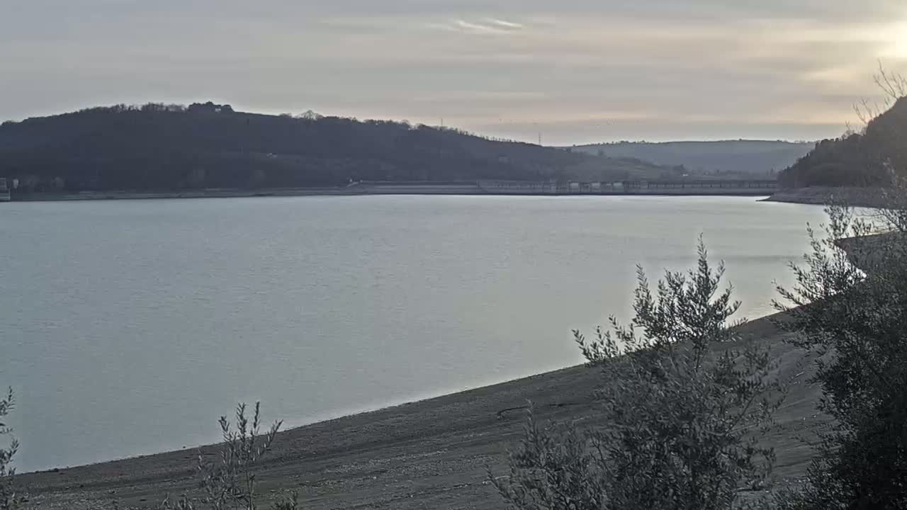 A calm, light-blue lake is partially bordered by a low, sandy shore under a pale, overcast sky, with dark hills in the background.