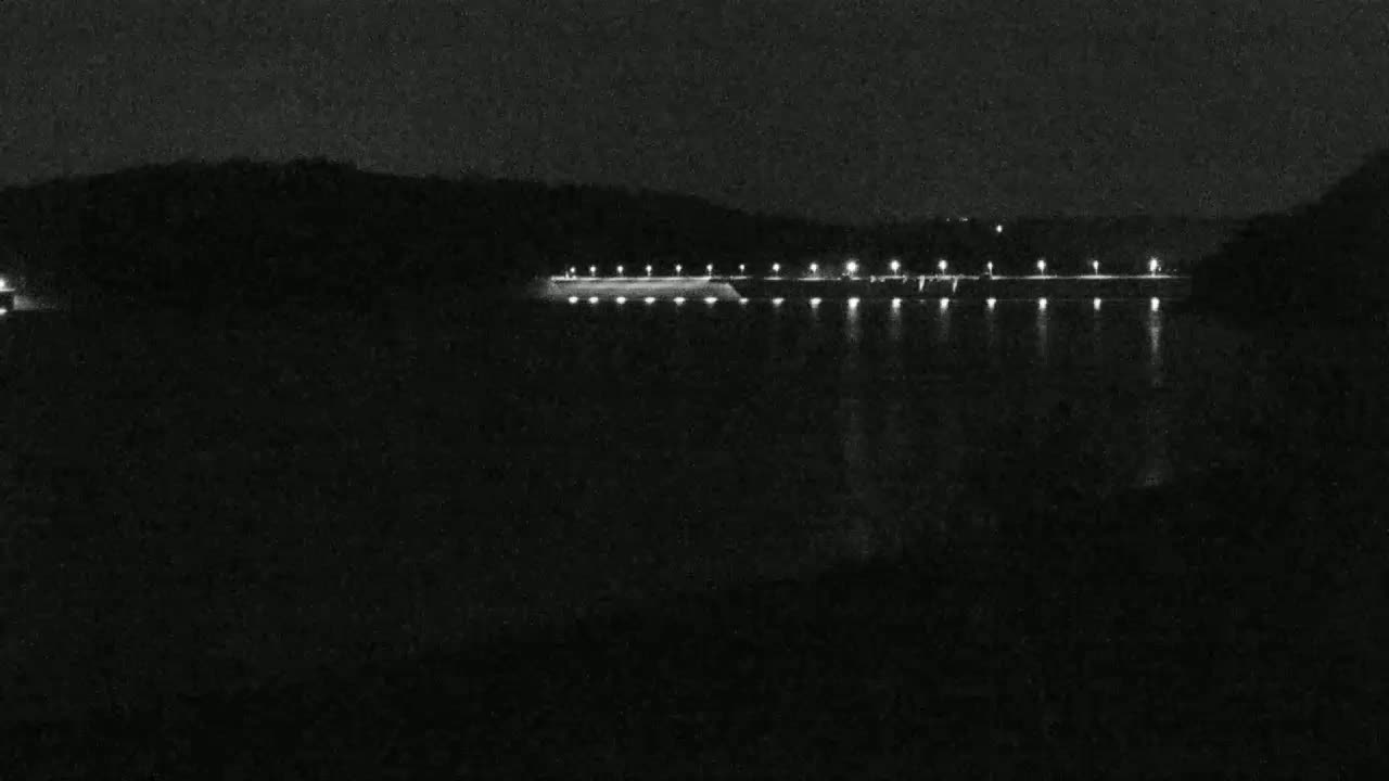 At night, a long, low structure with lights is visible across a dark body of water, with dark hills in the background.