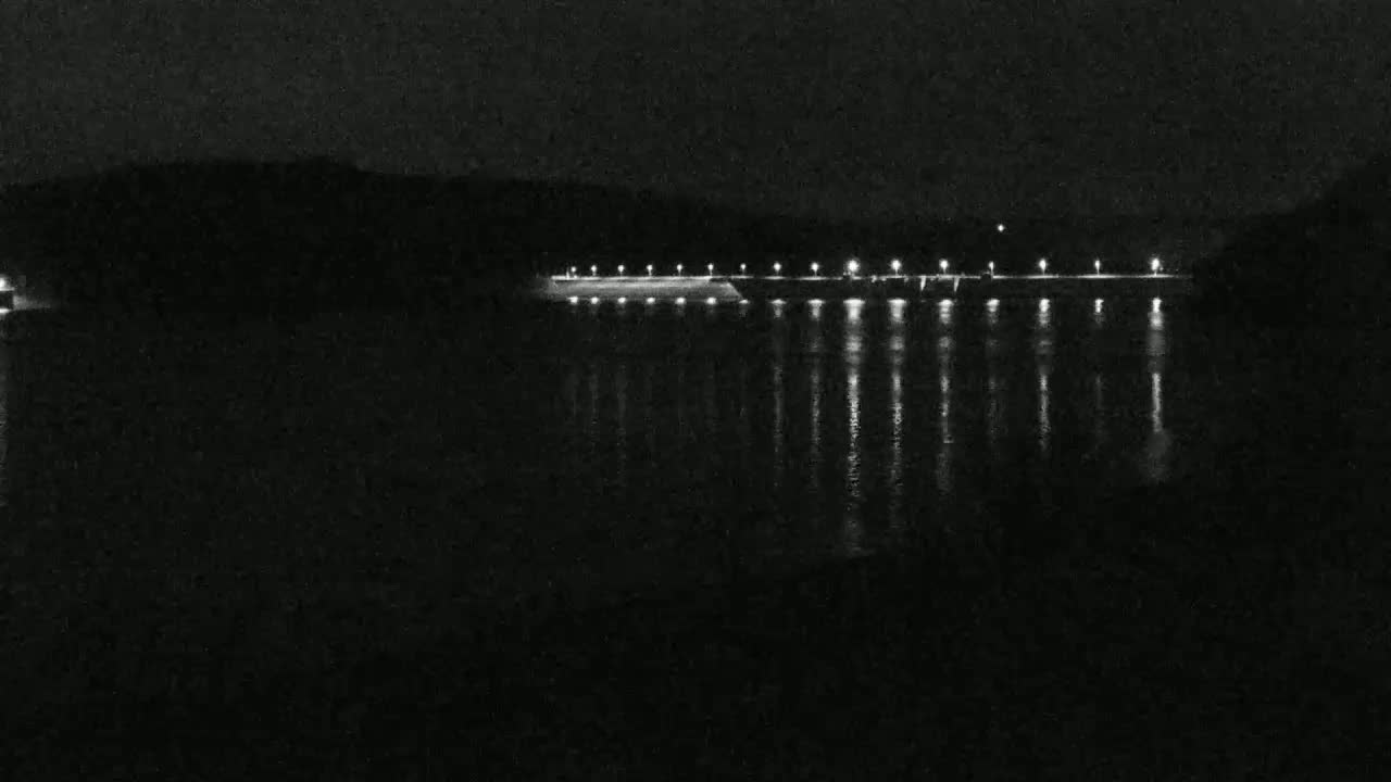 At night, a long, illuminated dam stretches across a dark body of water, reflecting the lights onto its surface.