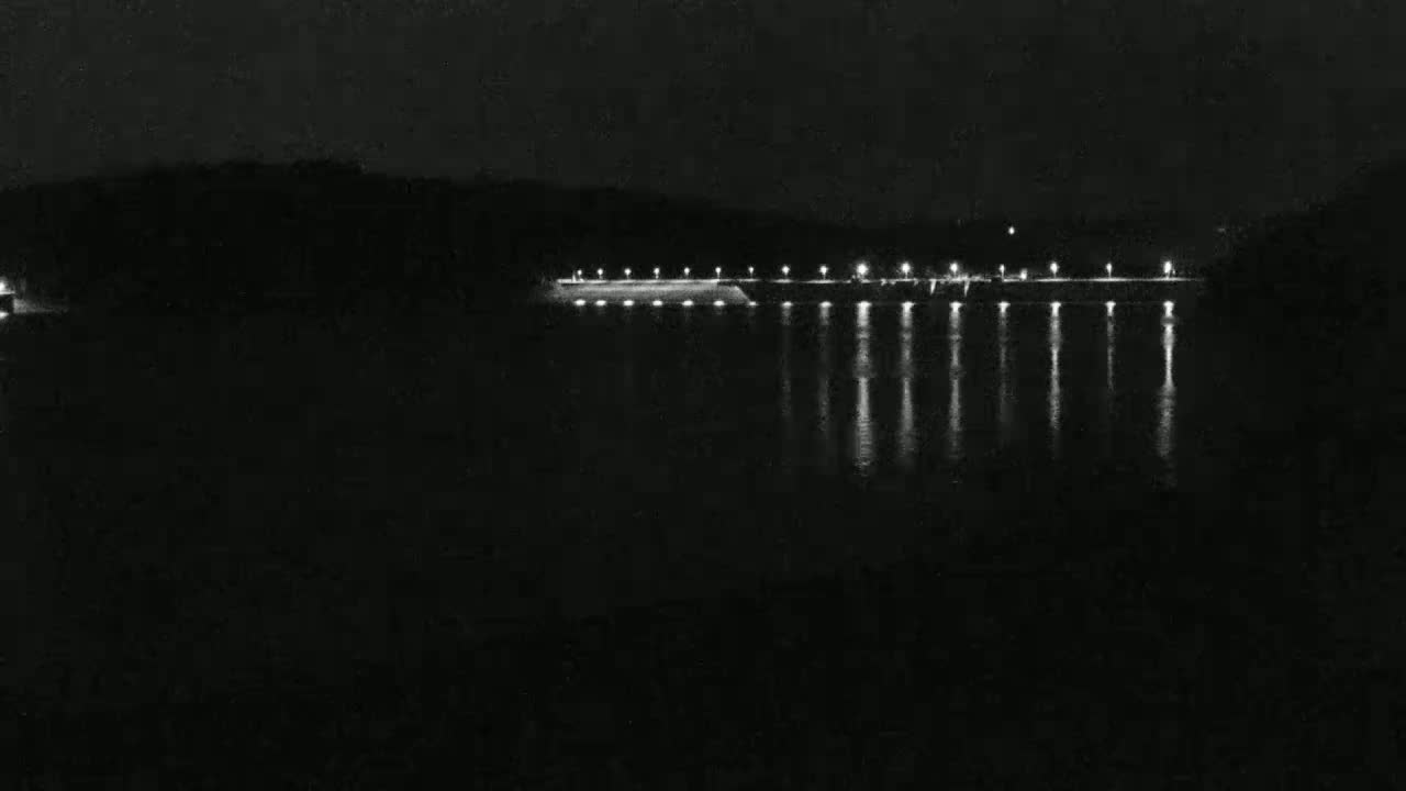 At night, a long, low structure with lights is illuminated along a dark body of water, reflecting the lights onto its surface.