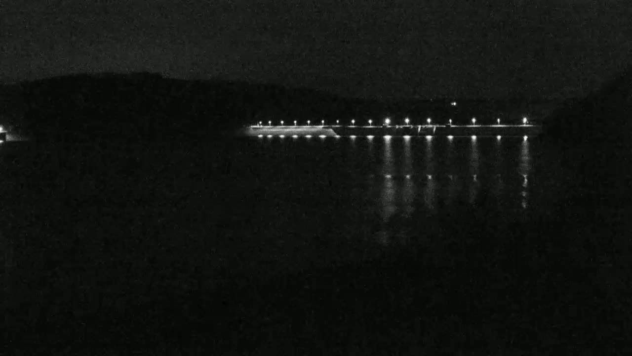 A long, dark structure with evenly spaced lights is illuminated at night, reflecting on a body of dark water.