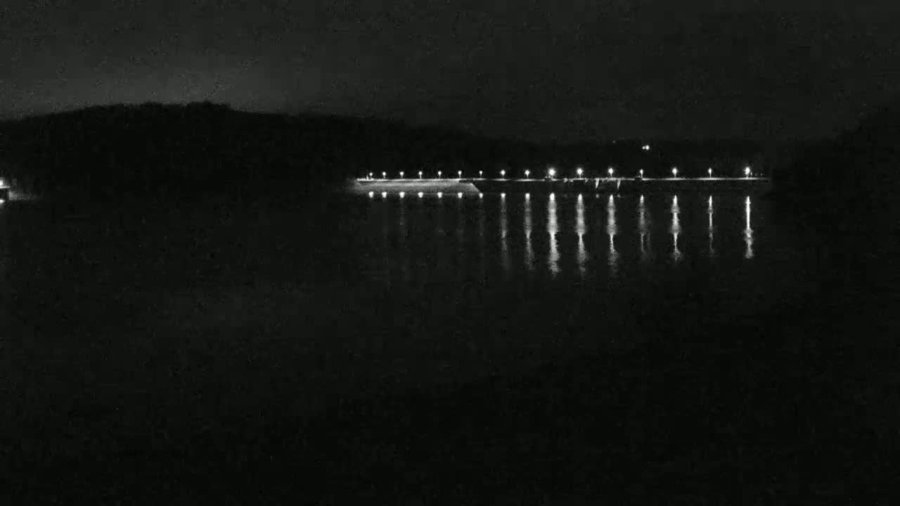 At night, a calm body of water reflects the lights of a structure on the far shore, set against a dark, wooded hillside.