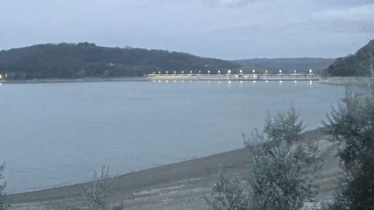 A calm lake with a low shoreline and a dam with lights visible in the distance under a dusky sky.