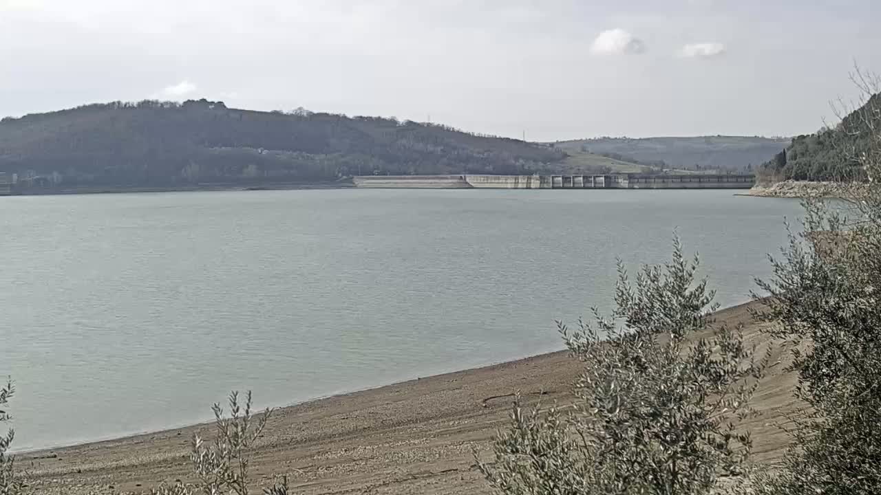A calm, light-green lake stretches out below a dam, with a receding shoreline revealing exposed sand and scrubby vegetation under a mostly clear sky.