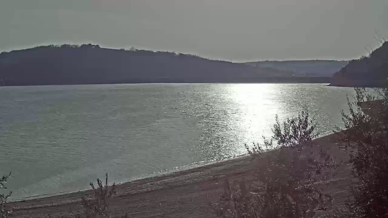 A calm body of water reflecting sunlight, with a sandy shore and hills in the background under a bright, mostly clear sky.