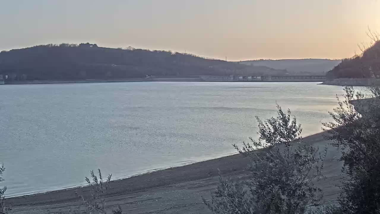 A calm body of water, possibly a lake or reservoir, stretches out under a pale sunset sky, with hills visible in the background and a sandy shoreline in the foreground.