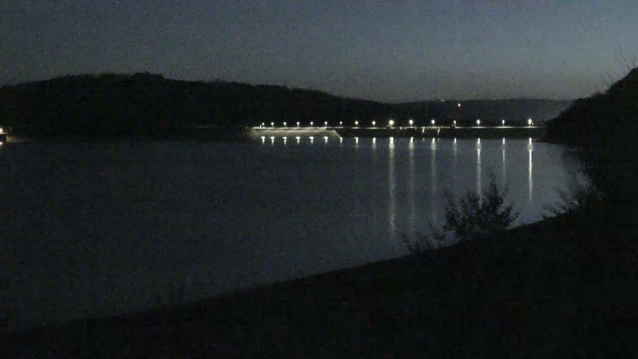 A dark, nighttime view of a calm lake reflecting the lights of a dam extending across its width, framed by hills in the background.