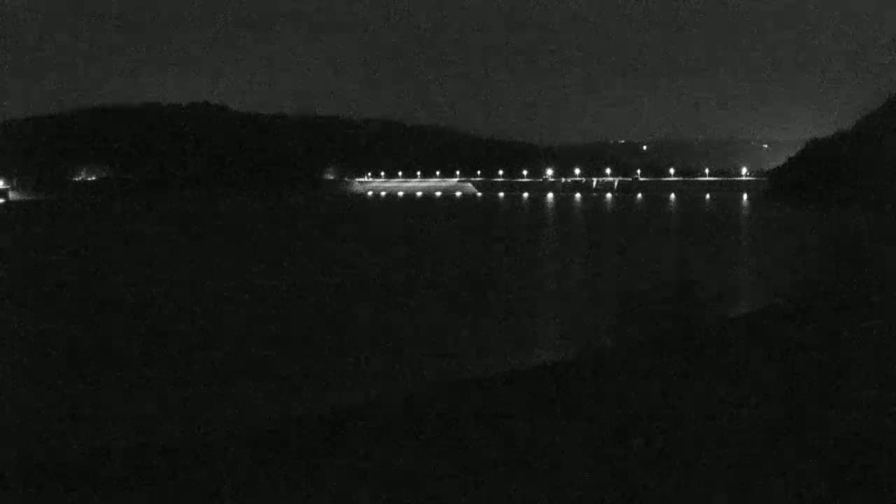 At night, a long, illuminated dam stretches across a dark body of water, backed by a silhouette of hills.