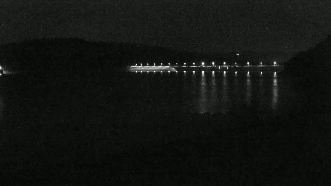 At night, a long, illuminated structure stretches across a dark body of water, flanked by dark land.