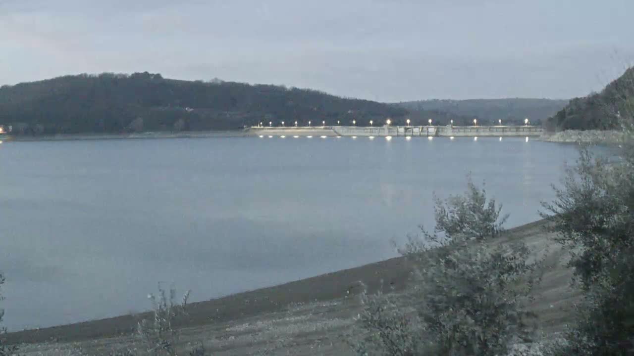 A calm lake at twilight reflects the lights of a dam against a backdrop of dark hills.