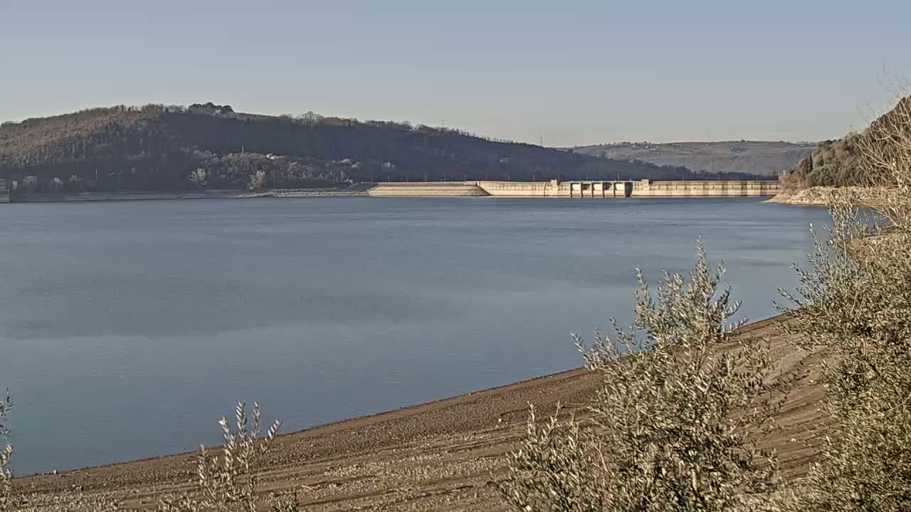 A calm lake stretches out before a dam, set against a backdrop of distant, tree-covered hills under a clear sky.