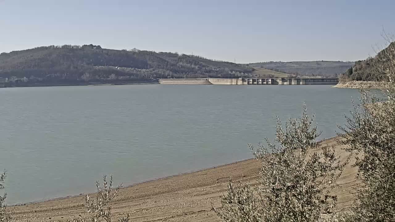 A calm lake with a dam in the distance, under a clear sky, is partially surrounded by bare, low hills and partially exposed sandy banks.