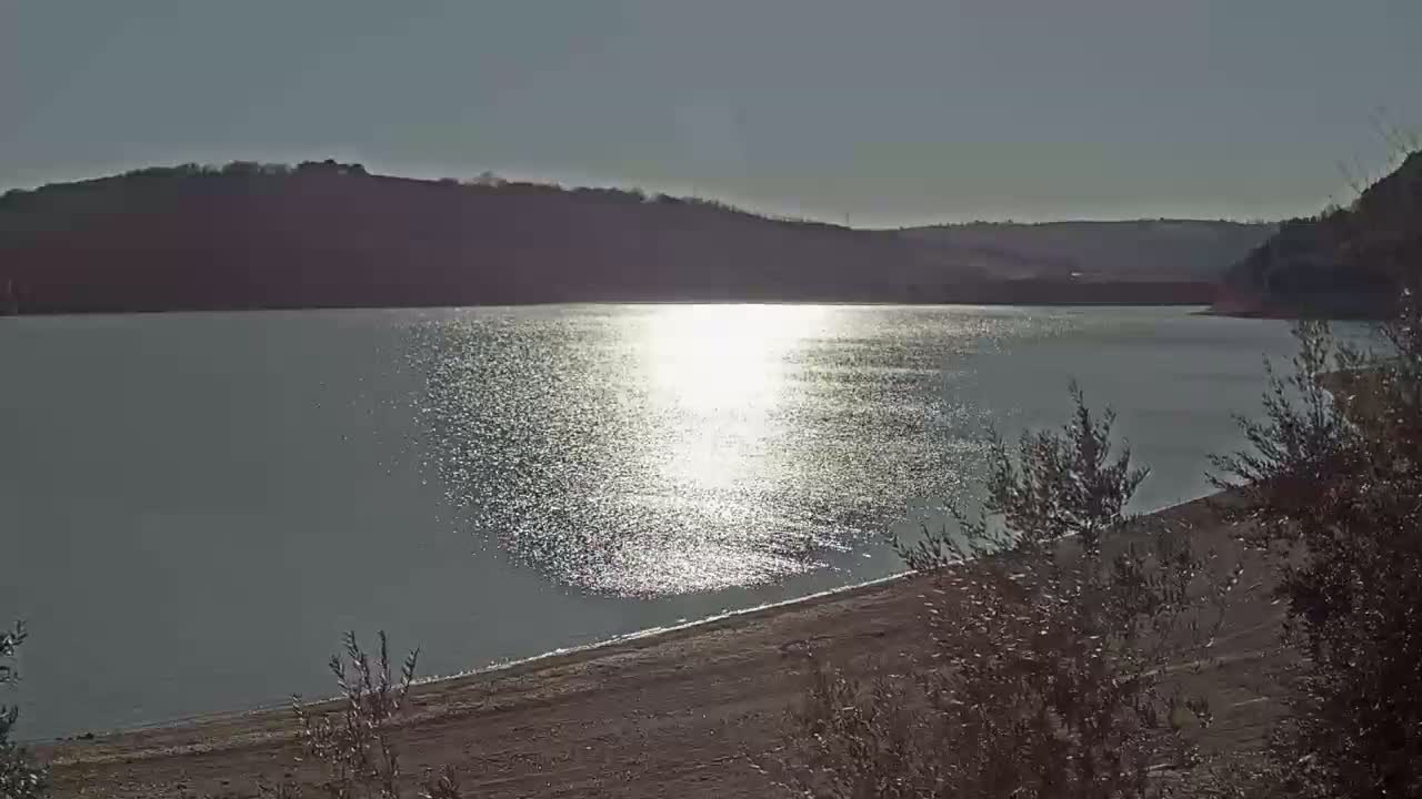A calm lake reflects the bright sun, its shoreline bordered by shrubs and hills under a clear sky.