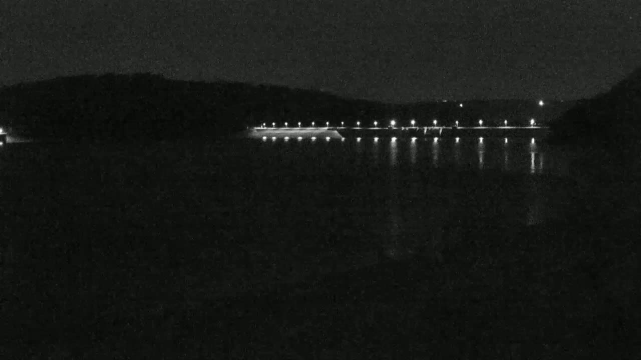A long, dark structure with lights is illuminated at night over a calm body of water, against a dark, hilly background.