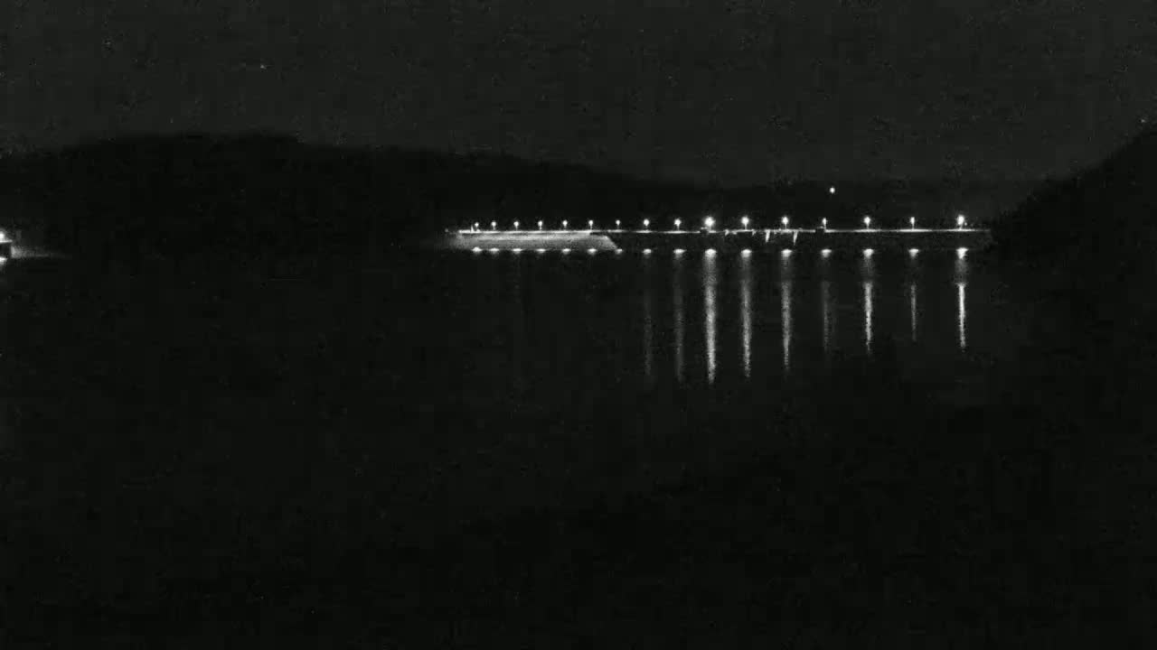 At night, a long, low structure with evenly spaced lights is reflected in a dark body of water.