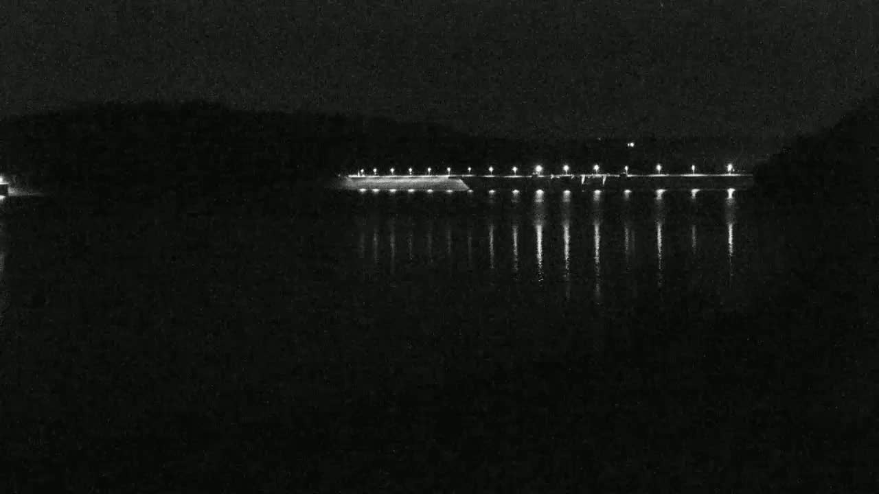 A dark, nighttime scene shows a long, illuminated structure stretching across a calm body of water, reflecting the lights.