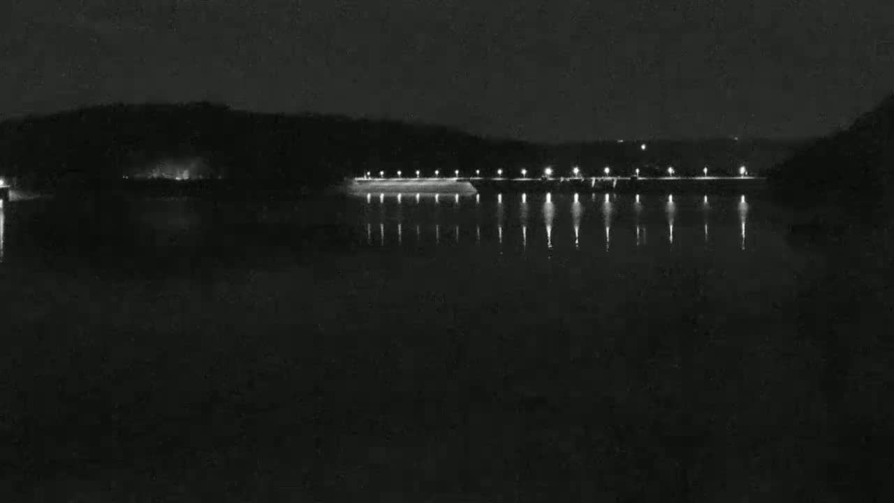 A nighttime view of a calm body of water reflecting the lights of a structure along its shore, which is backed by a dark, hilly landscape.