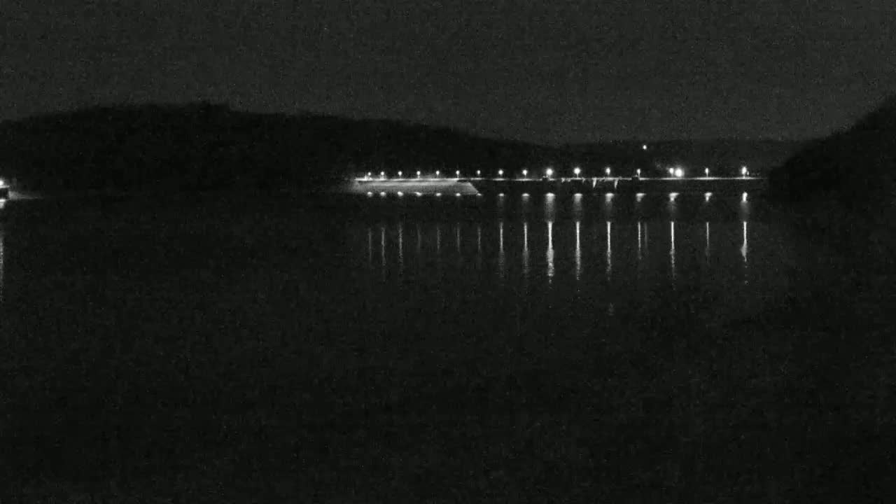 At night, a dam with lights illuminating its top is reflected in a dark body of water, surrounded by a dark shoreline.