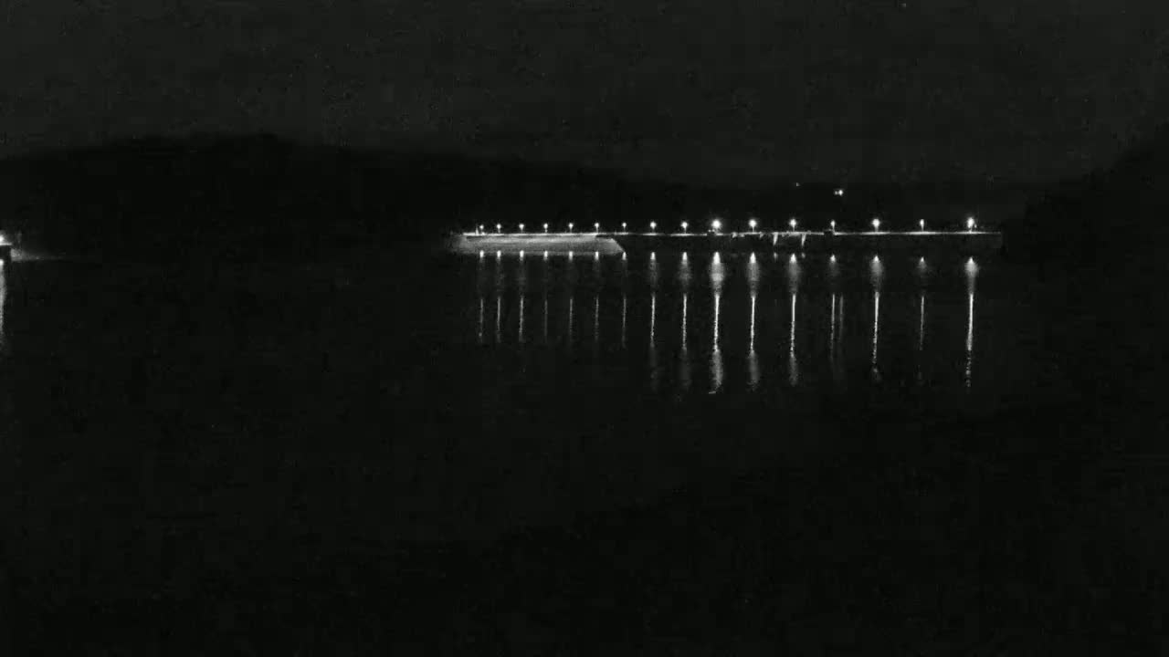 A dark, nighttime scene shows a long, lit pier extending over a calm body of water.