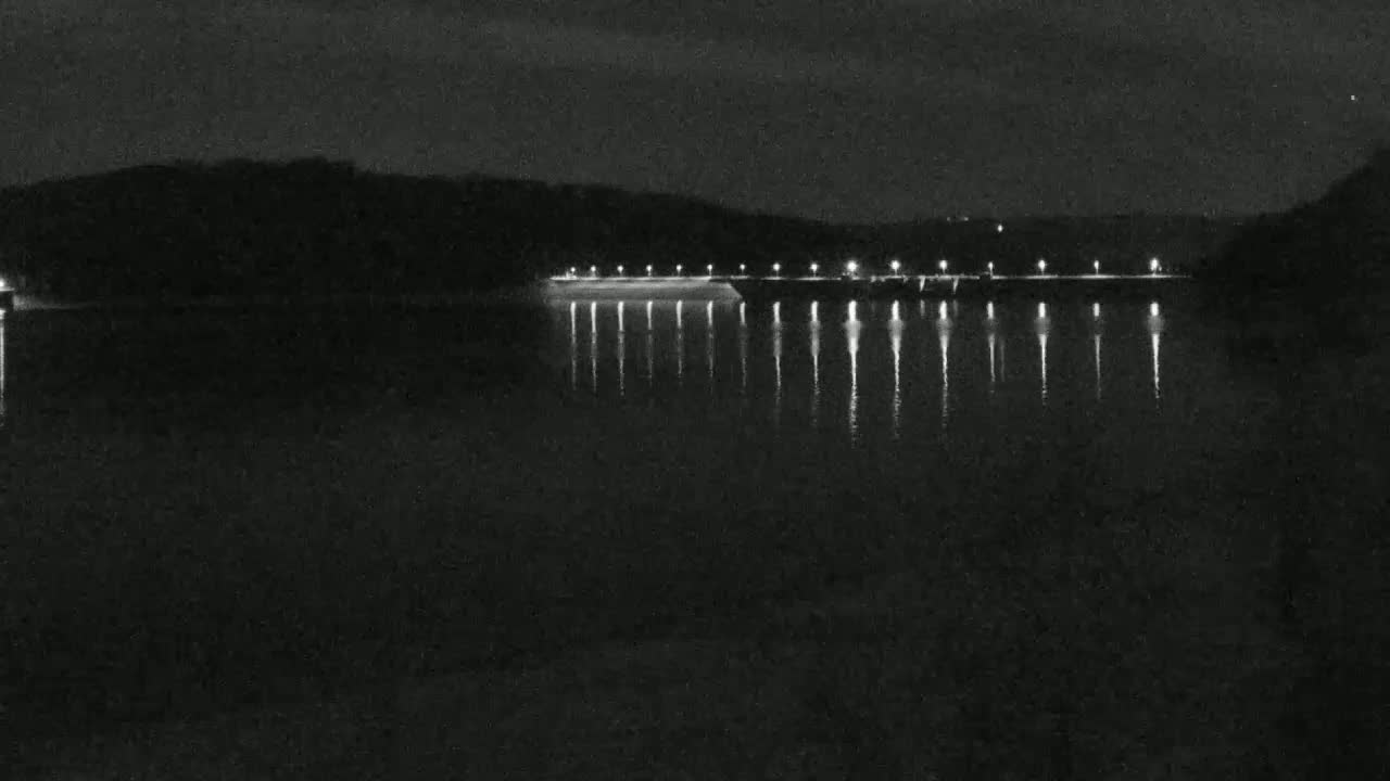At night, lights illuminate a long structure stretching across a calm body of water, reflecting the lights onto the dark surface.