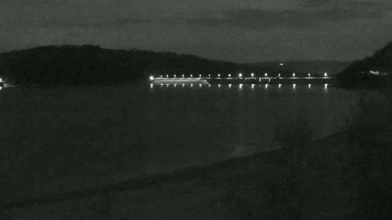 A nighttime view of a dam illuminated by lights across a dark body of water, with hills in the background under a dark sky.