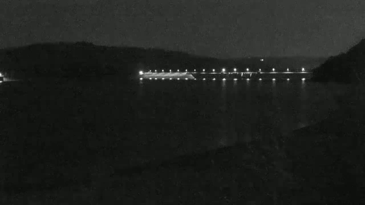 A dark, nighttime scene shows a lighted dam across a body of water, with hills visible in the background under a clear, dark sky.