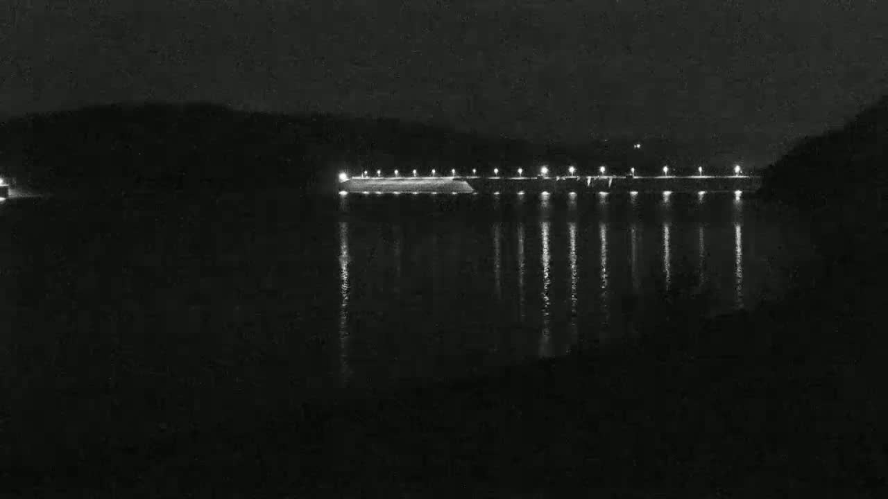 At night, a long, illuminated dam is visible across a dark body of water, reflecting the lights onto its surface.