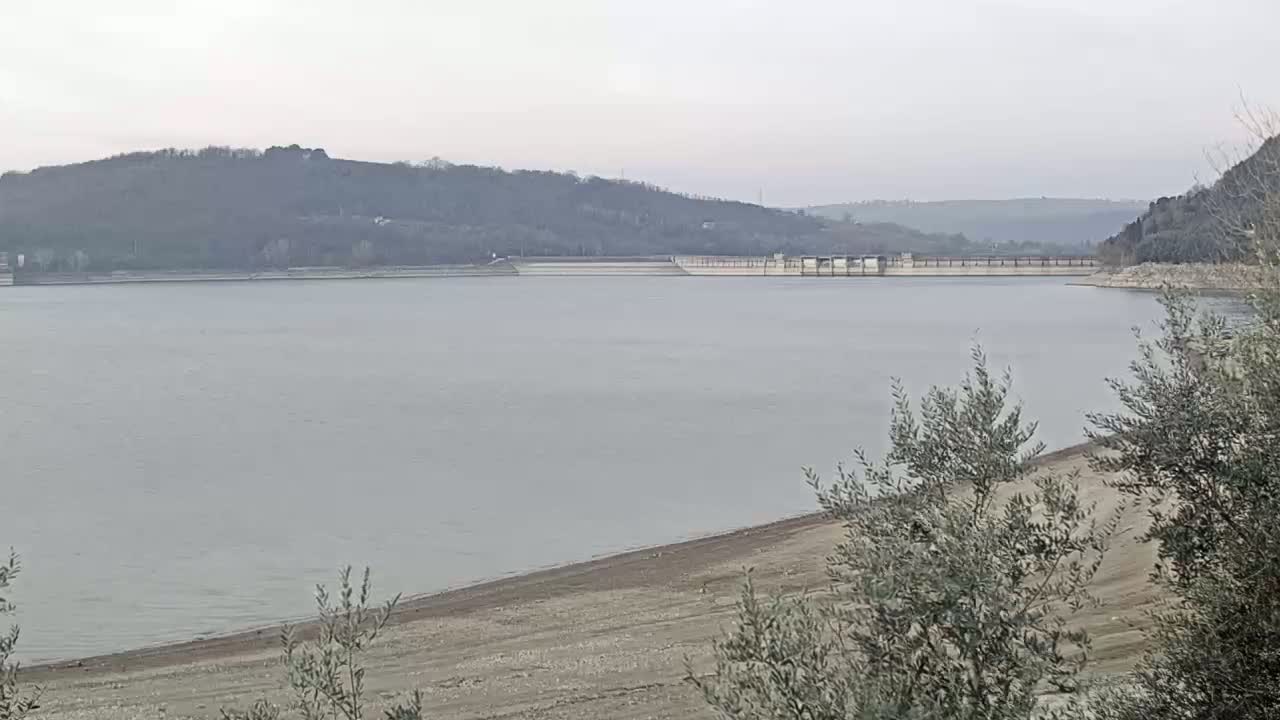 A calm, gray lake stretches out before a low, tree-covered hill, with a dam visible in the distance on an overcast day.
