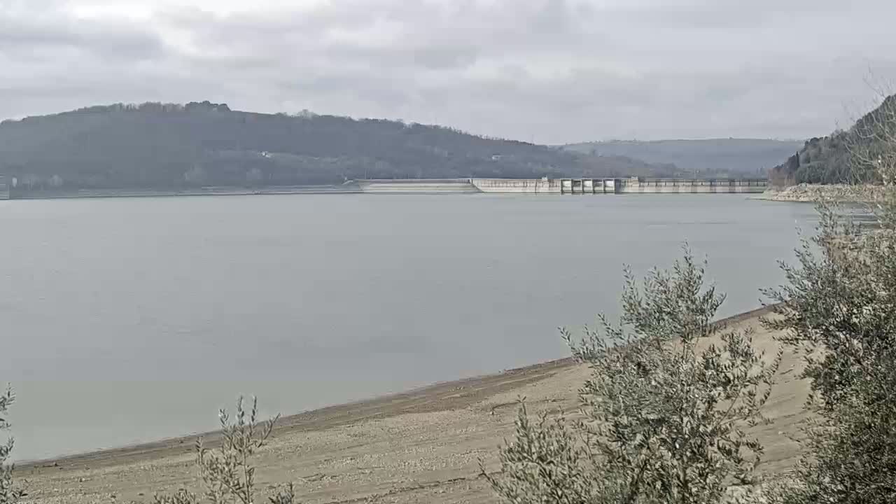 A large body of water with a dam in the distance, a low water level revealing a sandy shore, and overcast skies.