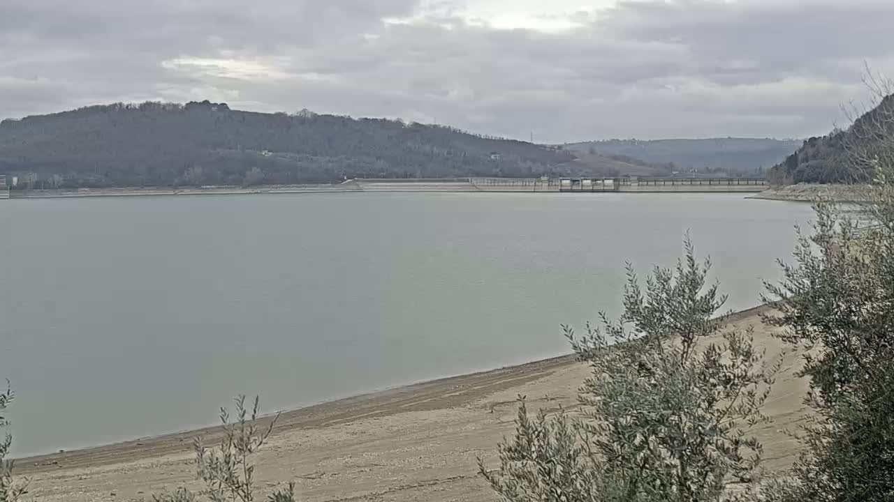 A calm, gray-toned lake with a low water level, exposing a sandy shoreline, is situated beneath a cloudy sky and hills covered in leafless trees.