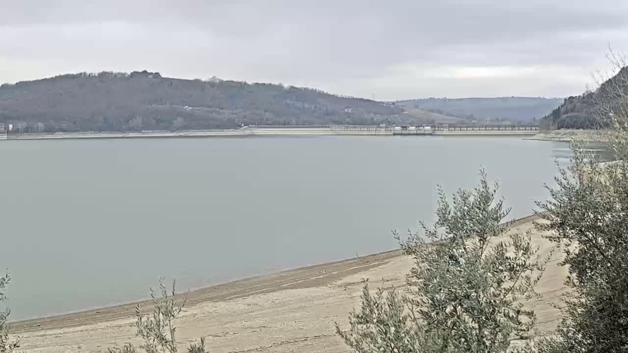 A calm, gray-water lake with a low water level exposing a sandy shoreline, is backed by hills covered in leafless trees under an overcast sky.