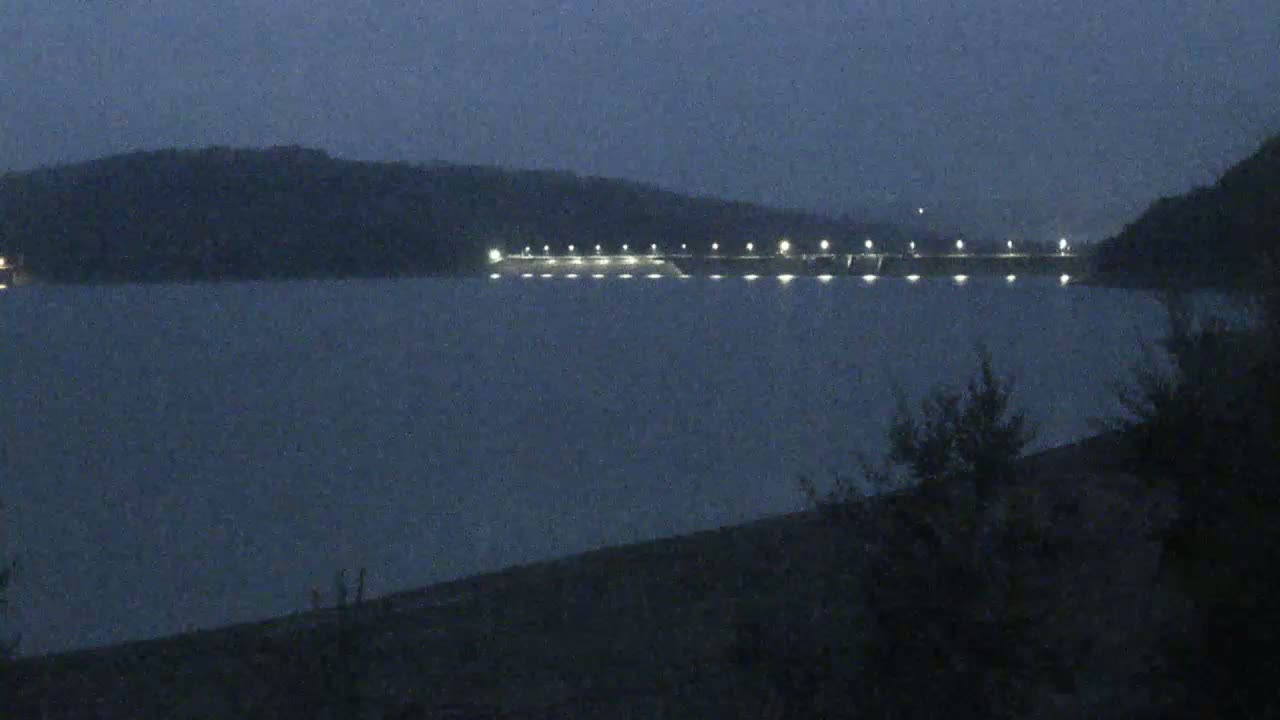 A long, illuminated dam is visible across a dark, calm body of water under a night sky.