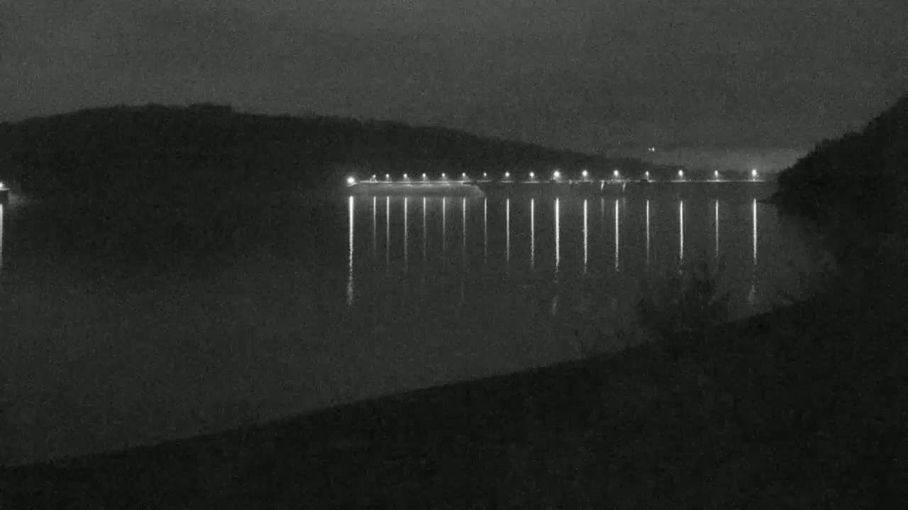 At night, a long, illuminated structure stretches across a dark body of water, reflecting light onto the still surface, with hills visible in the background under a dark sky.