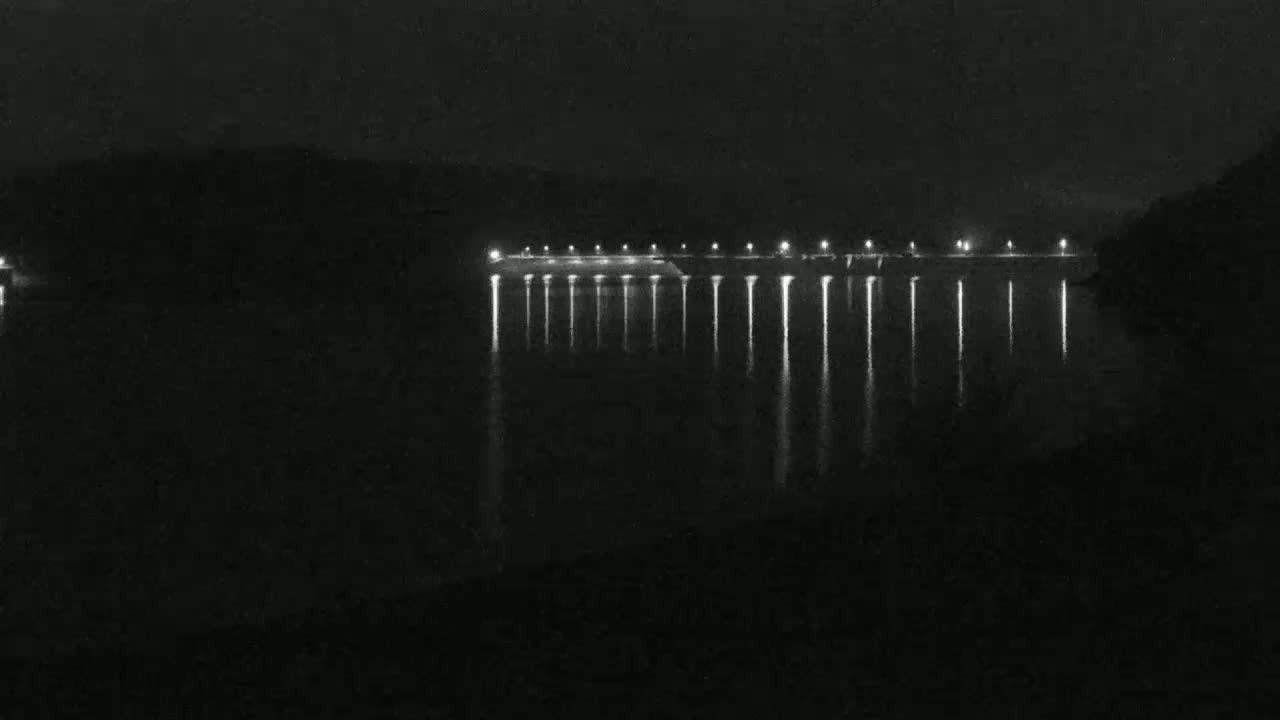 A long, dark structure with lights along its length is reflected in dark water at night.