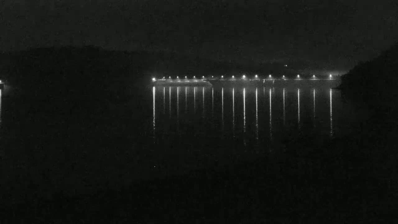 At night, a long, illuminated structure stretches across a dark body of water, its lights reflecting on the still surface.