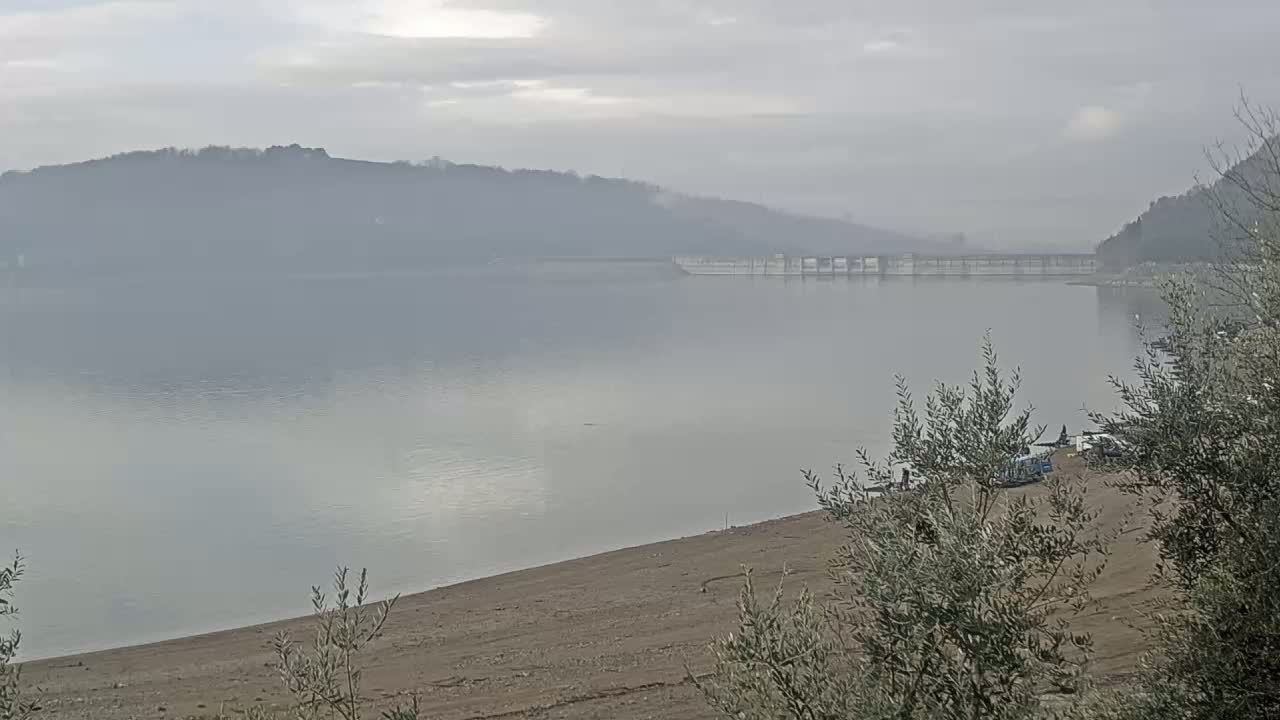 A calm, foggy lake stretches out to a distant dam nestled between hazy hills.
